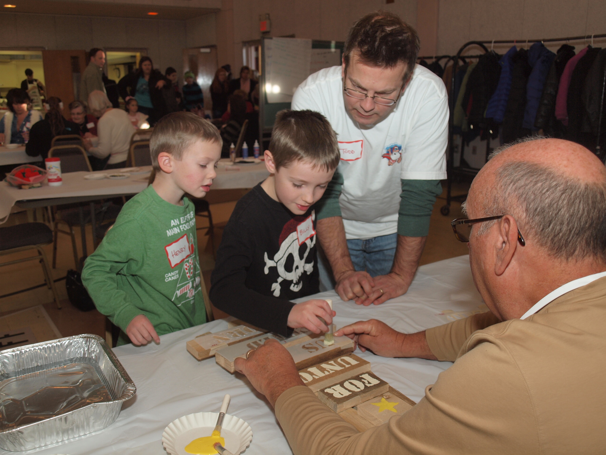 Henry Slater (left) watches closely as his brother Billy takes a turn at hand-stenciling a reclaimed-wood tree, with dad Todd Slater looking on. Volunteer Max Oehler (right) lends a steadying hand at last year’s Advent Festival.